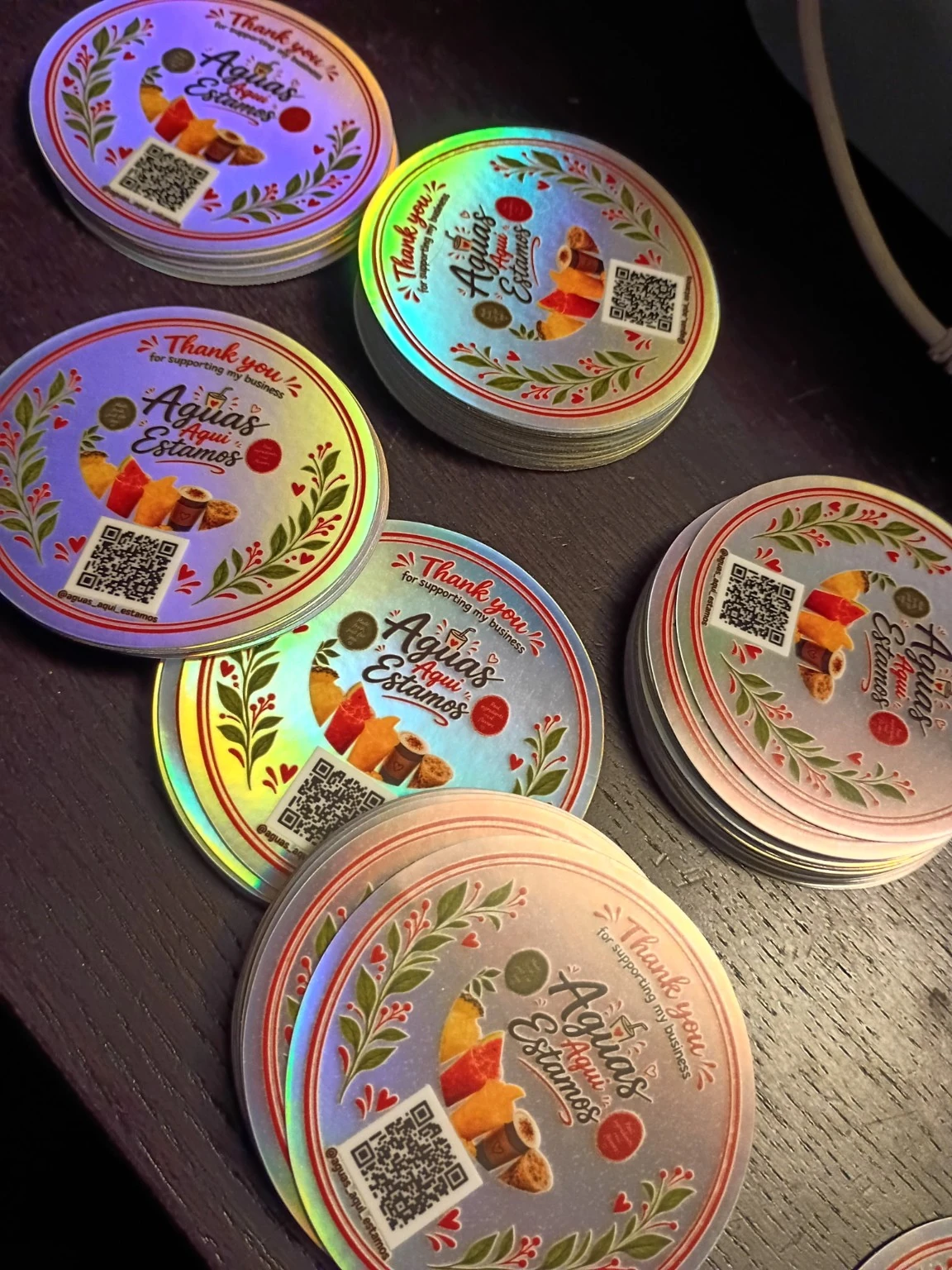Stacks of finished Aguas Aqui Estamos round holographic logo and Instagram QR code stickers on a dark wooden table.