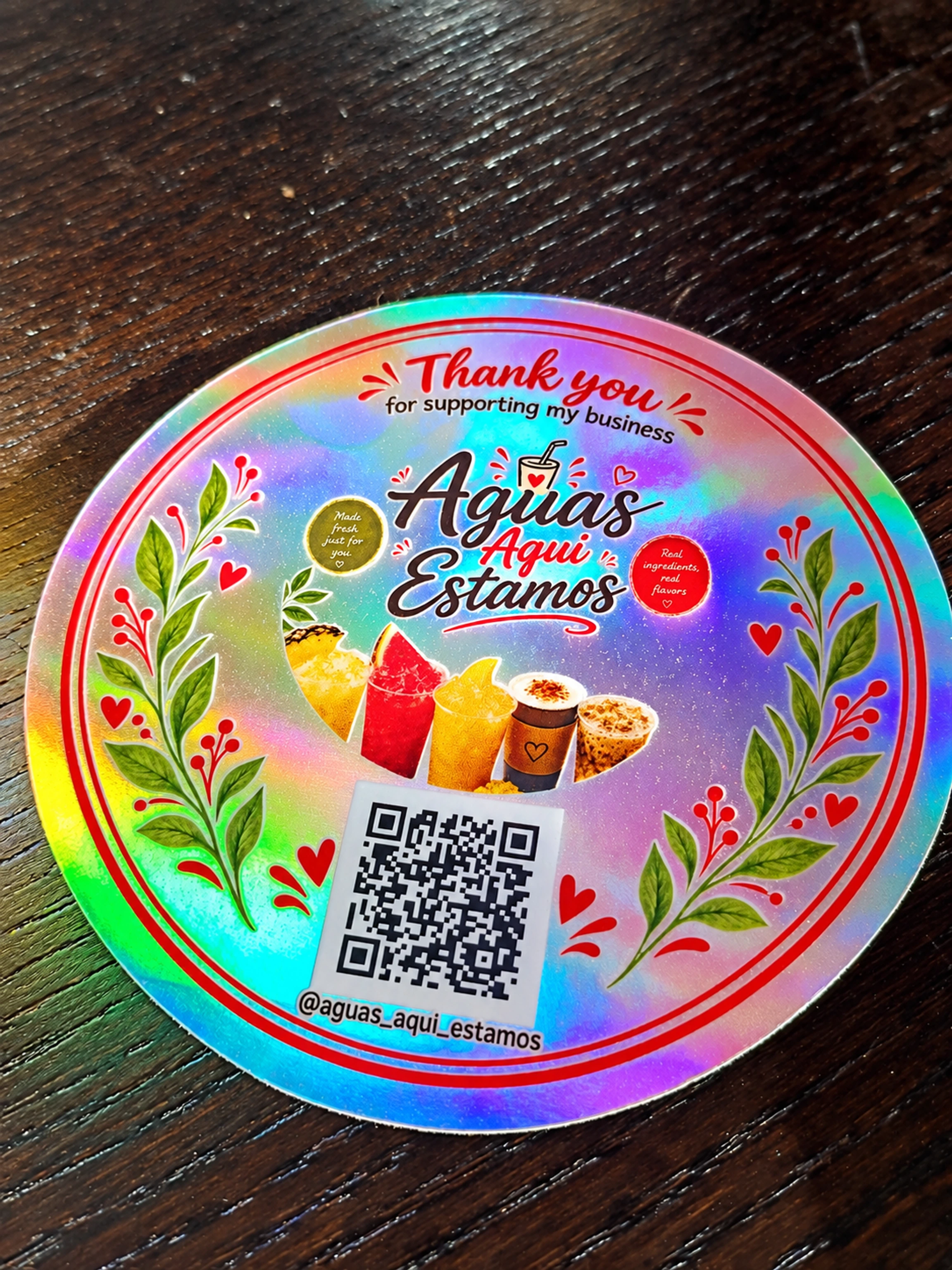 A round Aguas Aqui Estamos holographic logo sticker with colorful drink illustrations, leafy borders, red hearts, and a scannable Instagram QR code on a dark wooden table.